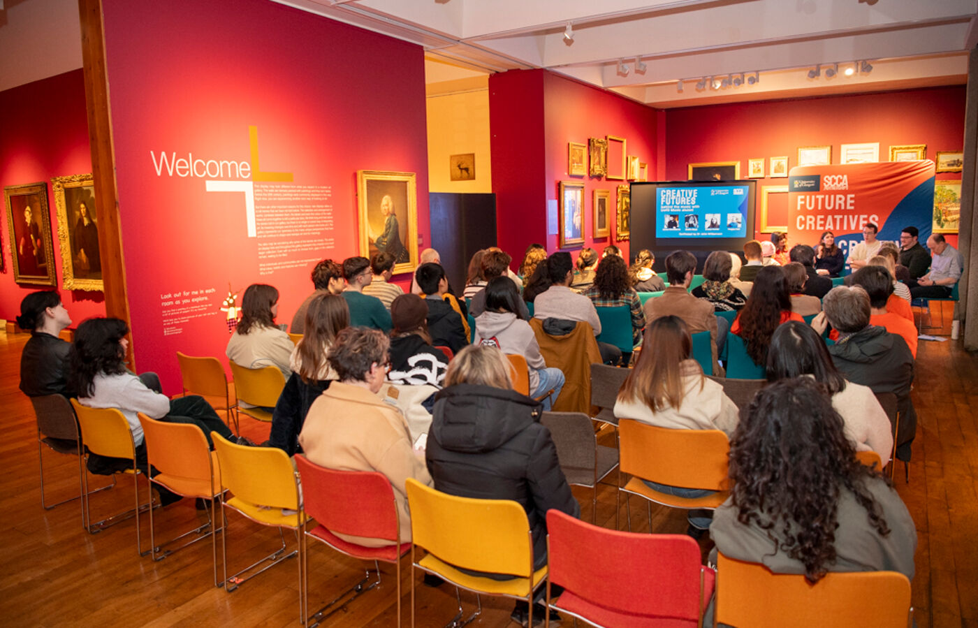 An audience is seated in a gallery with colorful chairs, watching a presentation with the 