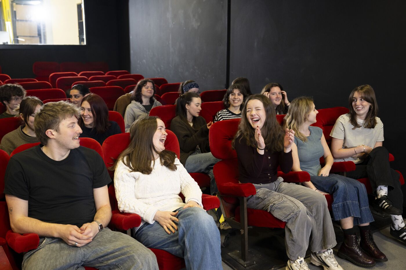 A group of young people are laughing while seated in red cinema chairs.