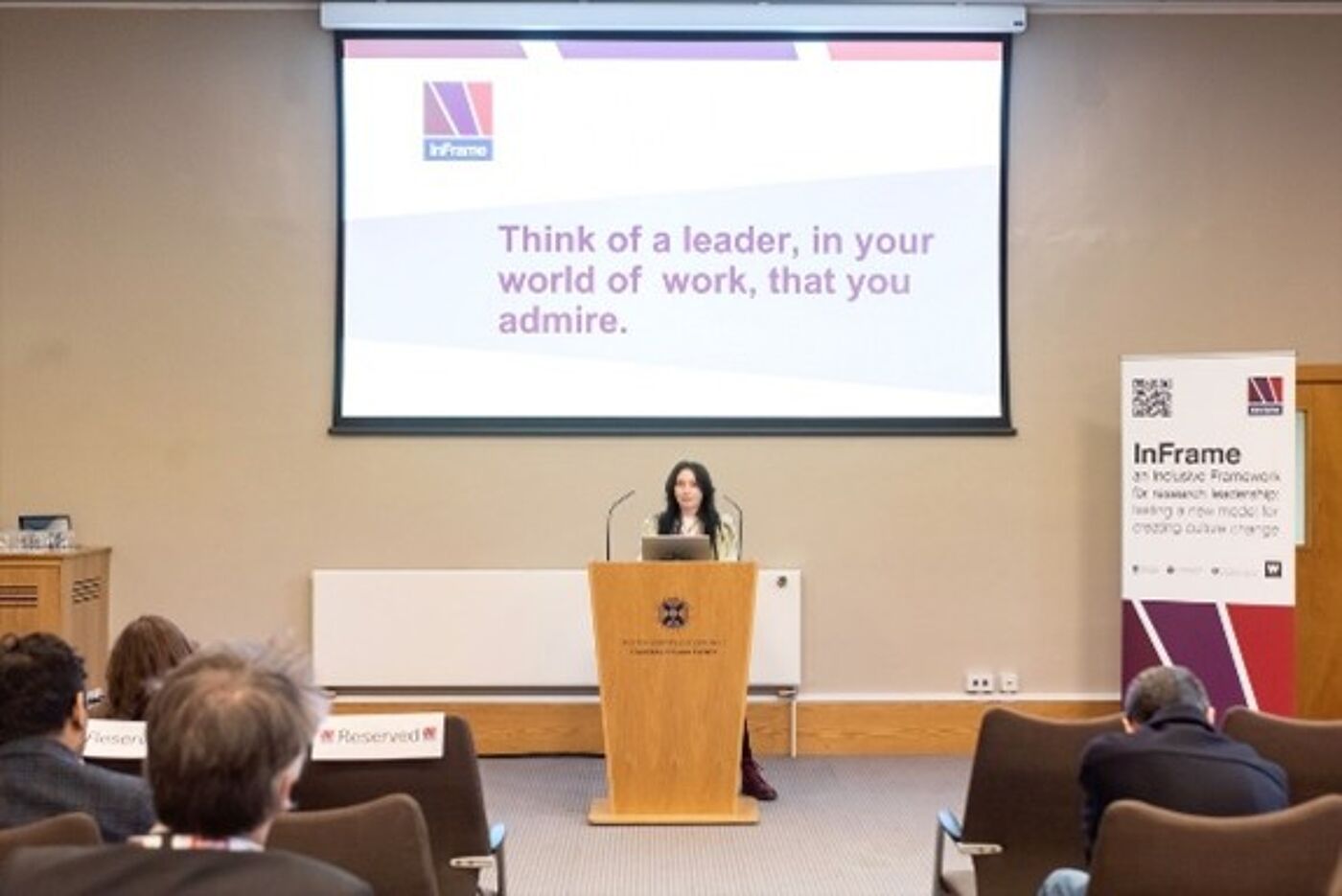 Audience seated in a lecture auditorium facing a stage where a speaker stands at a podium. Behind them, a large screen displays the text: “Think of a leader, in your world of work, that you admire.” InFrame banners and logos are visible, indicating a research culture or leadership event.
