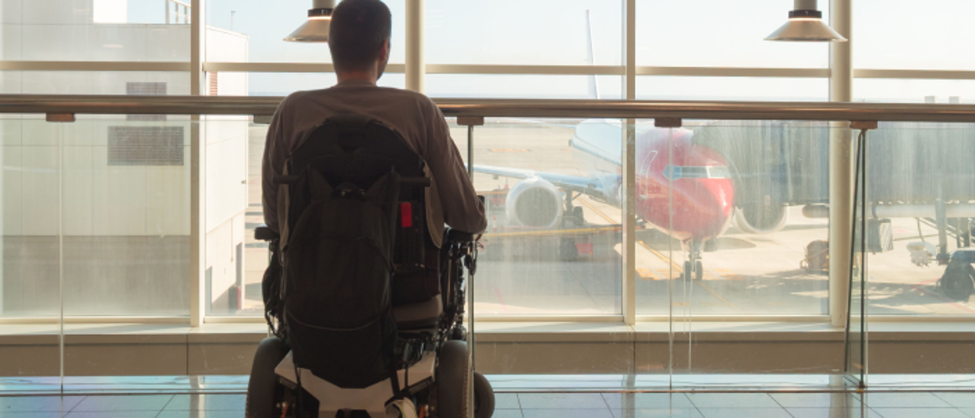 Person with disability using wheelchair observes the airport scene, waiting near the departure gate. The bright terminal features windows showing airplanes outside.