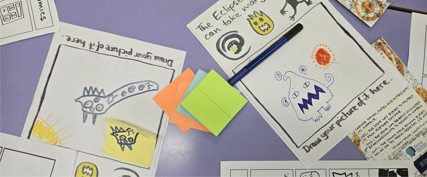 A photo of a school desk with paper and post-it notes spread out. The paper shows children's drawings of monsters.