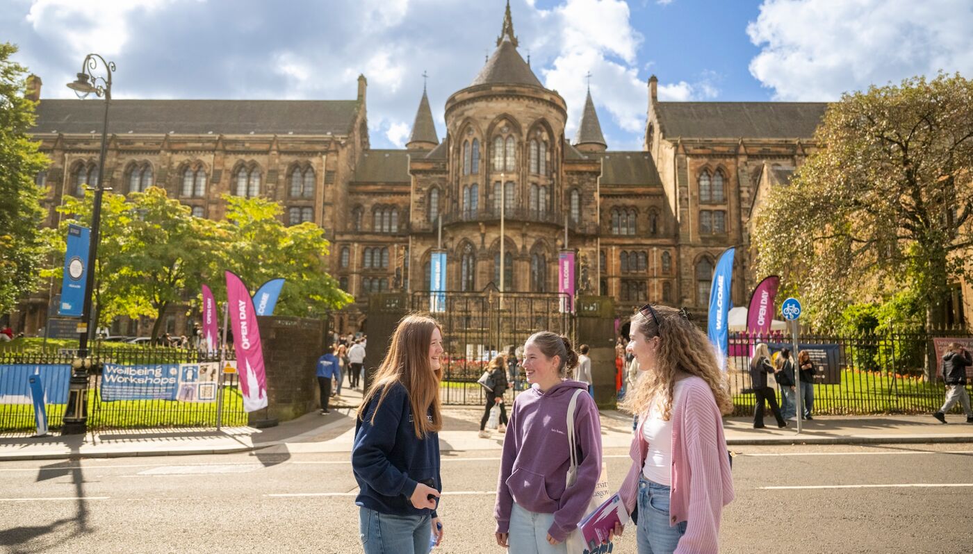 Three students standing in front of the memorial gates at the University of Glasgow, with Open Day branding in the background