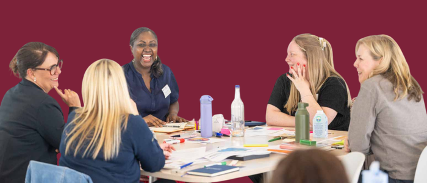 female leaders around a table knowledge sharing while looking engaged and happy at an executive education programme on a solid burgundy background