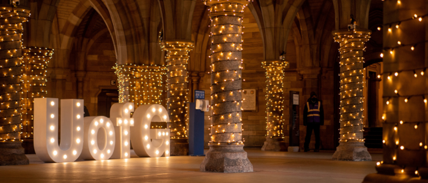 white letters spelling 'UofG' sitting in the cloisters in the evening with fairy lights lighting the space