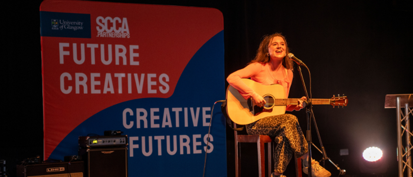 A student sits on a chair playing guitar in front of a large fabric banner including 'FUTURE CREATIVES, CREATIVE FUTURES' on an orange and blue background
