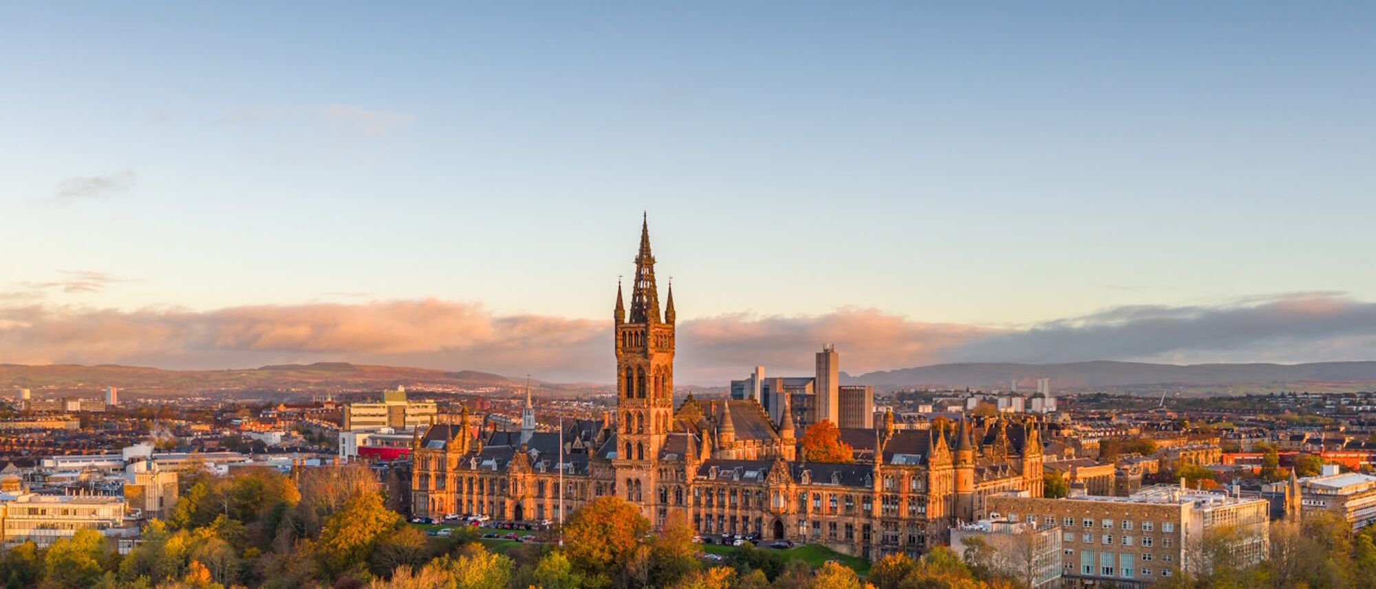 A drone view of the Gilbert Scott Building in autumn, reflecting red light at sunset, taken from the south looking north west, with hills in the background.