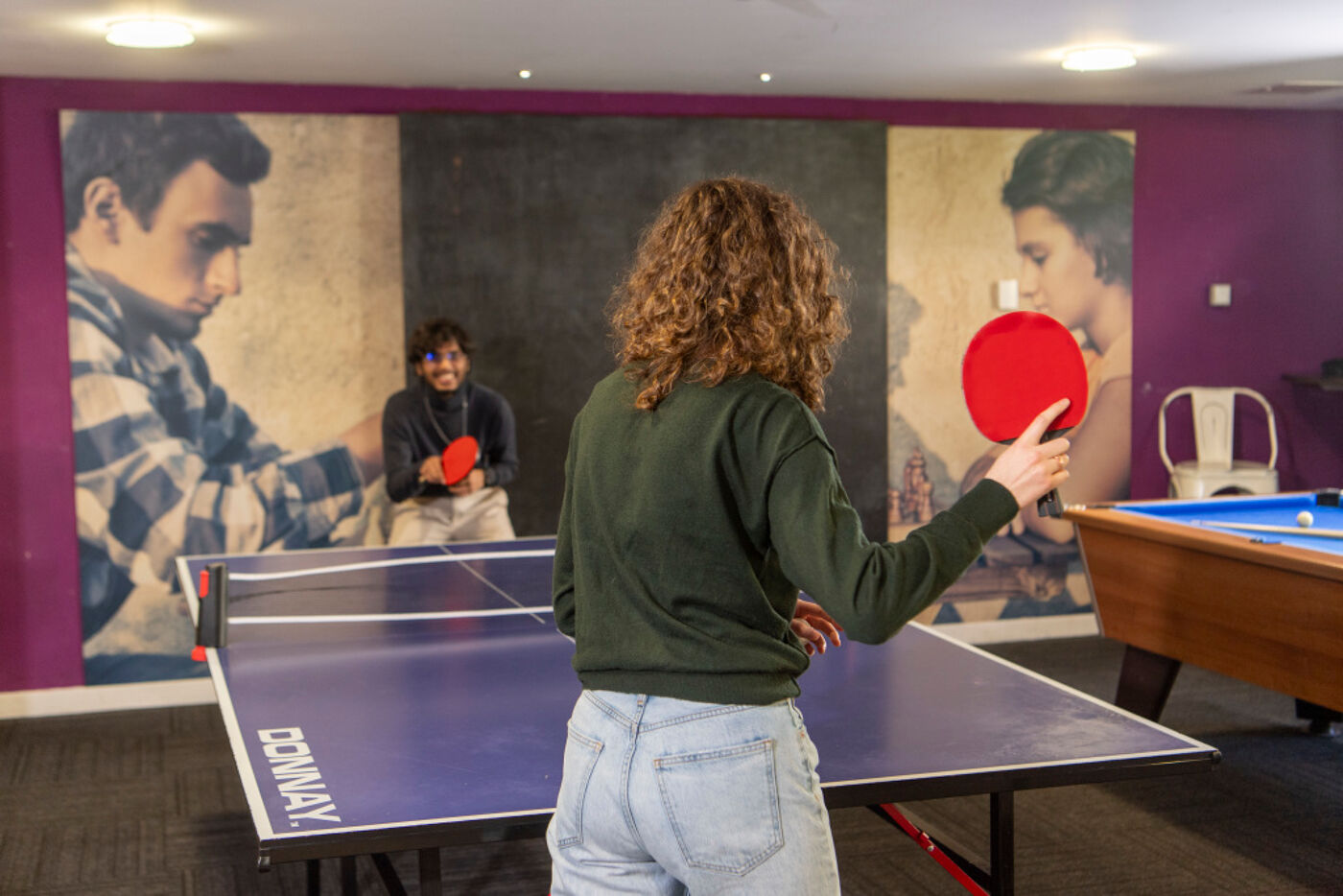 Students playing ping pong. The person facing us is smiling.