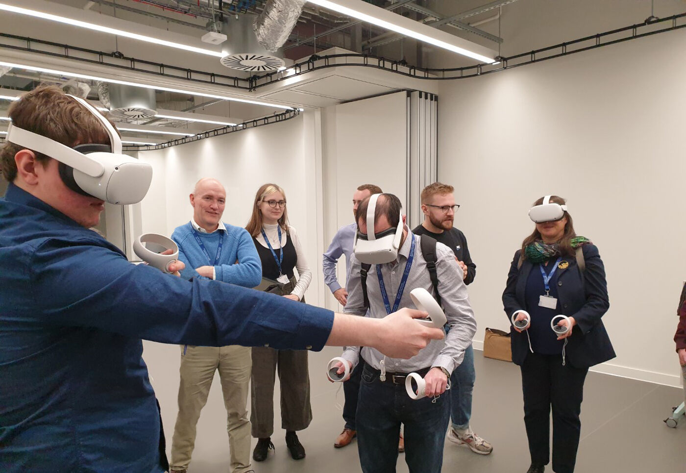 A group of people in a large room, so of them wearing VR headsets and stretching their arms out, holding small control devices