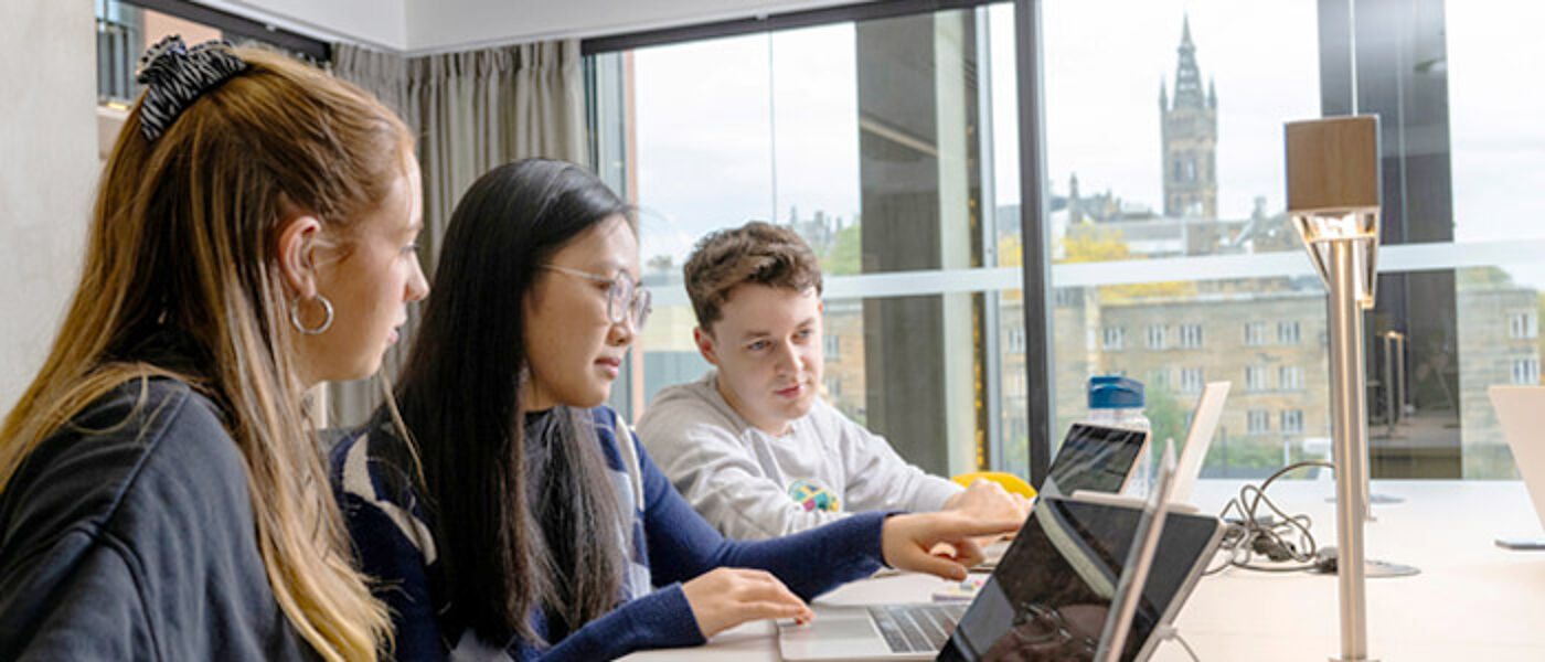 3 students in a study room working together sharing laptop screens with the historic Glasgow University Gilbert Scott Building Tower seen through the window in the background.