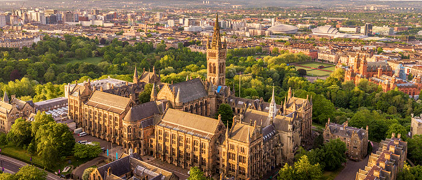 Glasgow University historic Gilbert Scott building, often nicknamed Hogwarts, immediately surrounded by greenery, with other historic and modern buildings in the background.