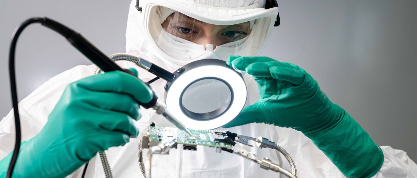 A researcher in a white protective suit is working on a delicate piece of equipment under a magnifying lens.