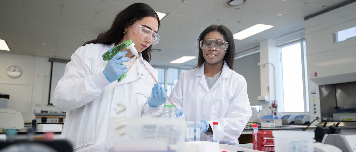 Two women in lab coats and safety glasses using a large pipette in a lab.