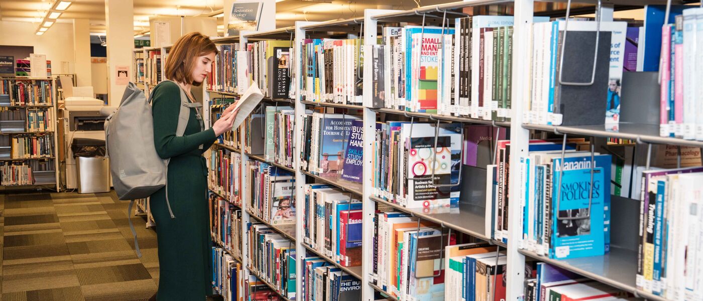 Woman in a green dress reading a book while standing in front of a set of bookshelves filled with books.