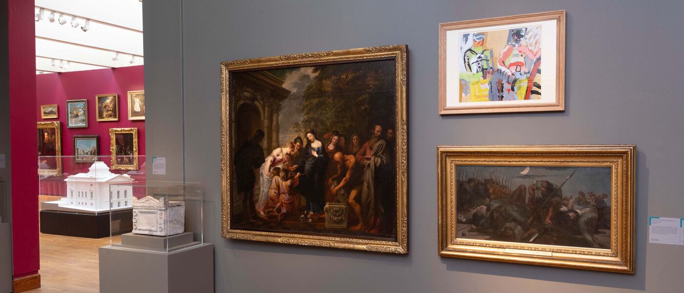 Three paintings hung on a grey wall, with a carved artefact in a glass case beside them