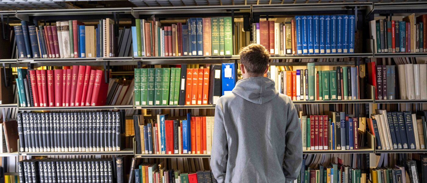 Person in a grey hoodie stands in front of a set of library shelves filled with books.