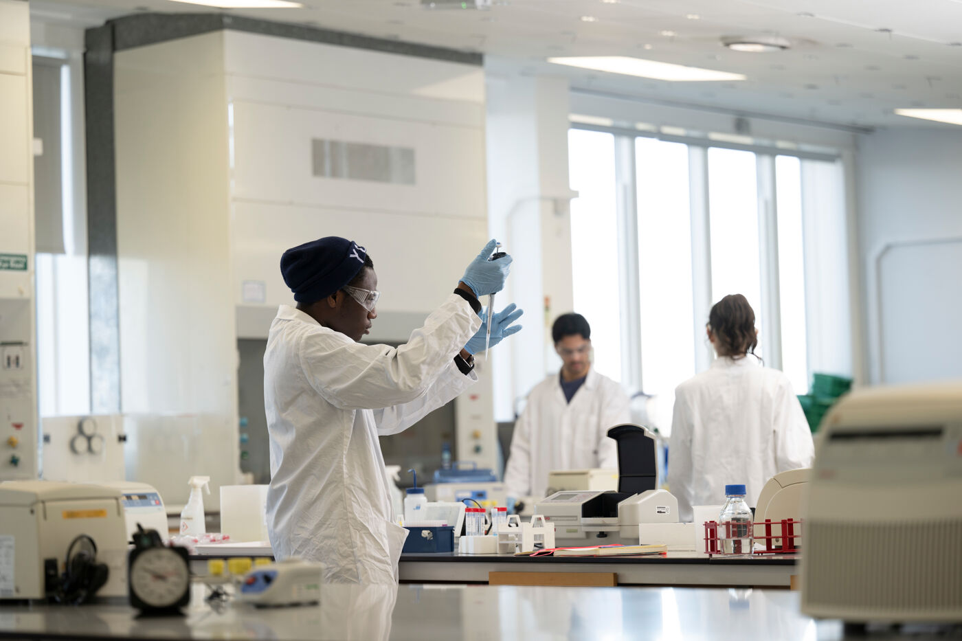 Student in a science lab, thy are wearing a white lab coat and using a pipette.