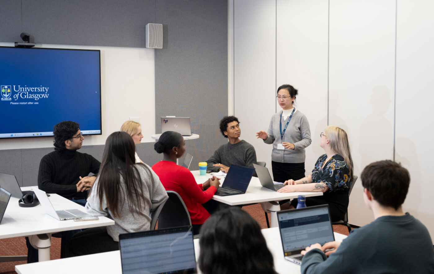A classroom setting with students sitting at multiple desks. They are listening to a staff member who is standing up and speaking.