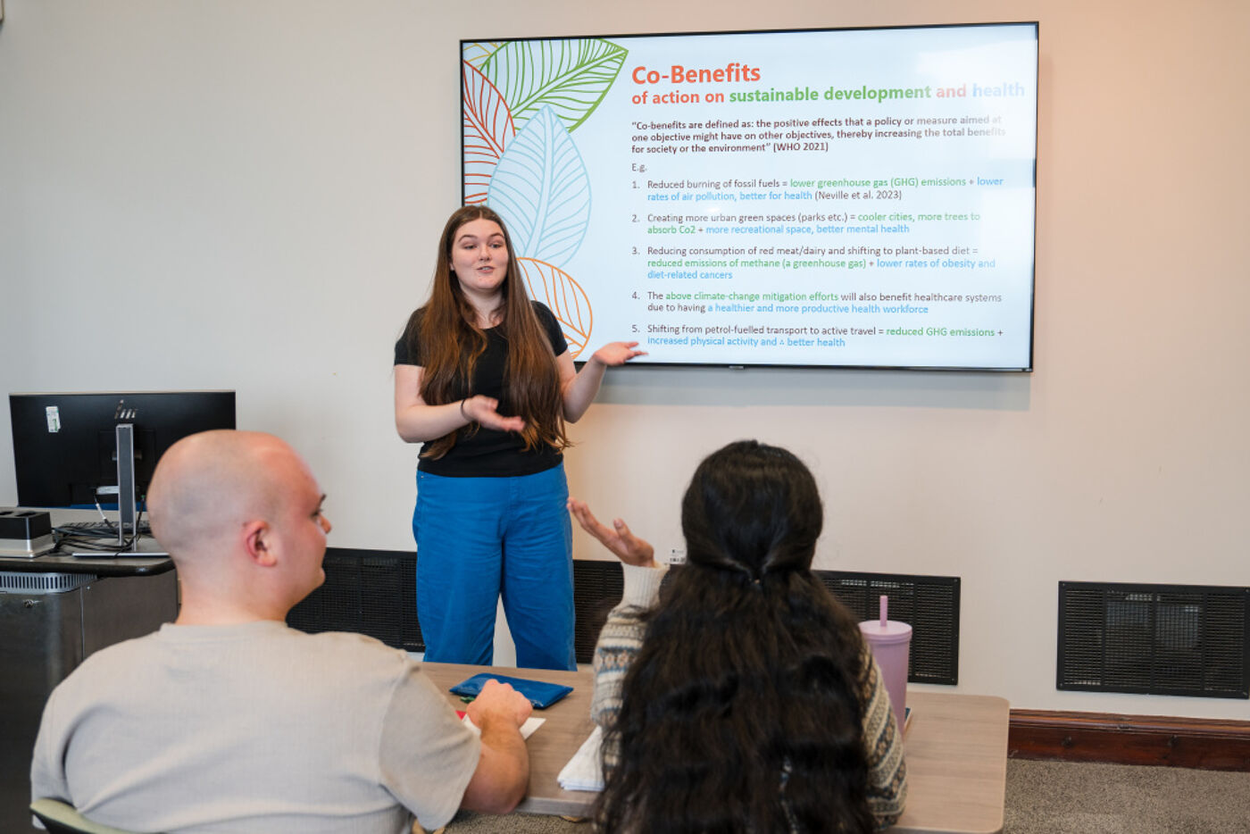 A student standing at the front of a class. They are gesturing and slides are on the projector behind them.
