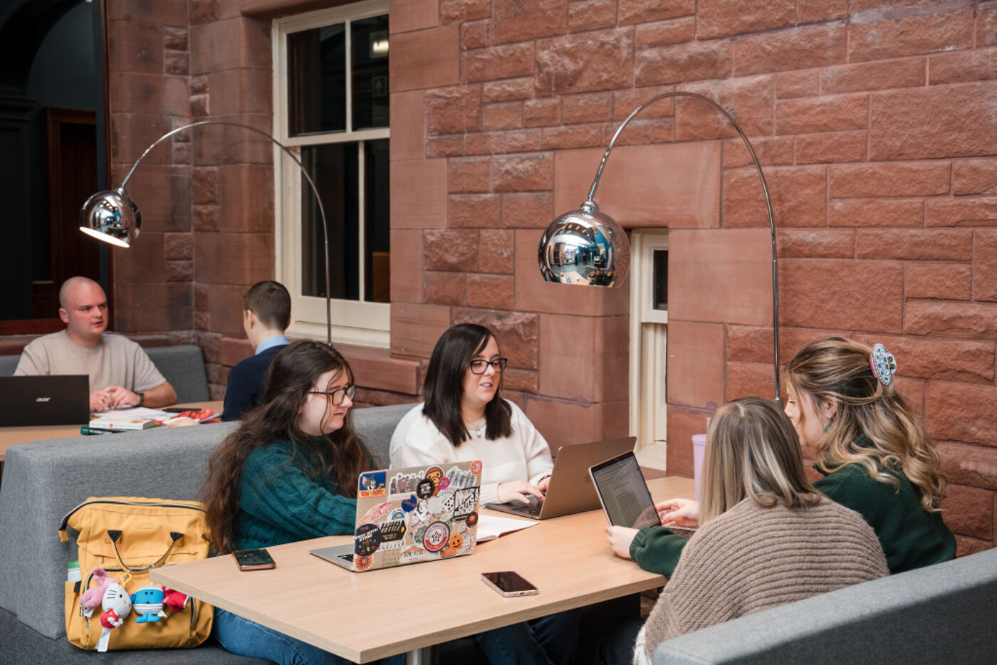 Four students sit together at a table. Two have their laptops out and they all are looking at the laptops and appear to be in discussion.