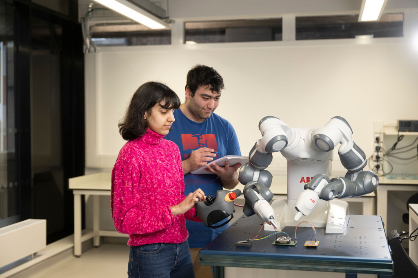 Two students stand next to a robot on a table. The robot has two arms and is manipulating an electronic circuit. One student has a controller and the other is taking notes.