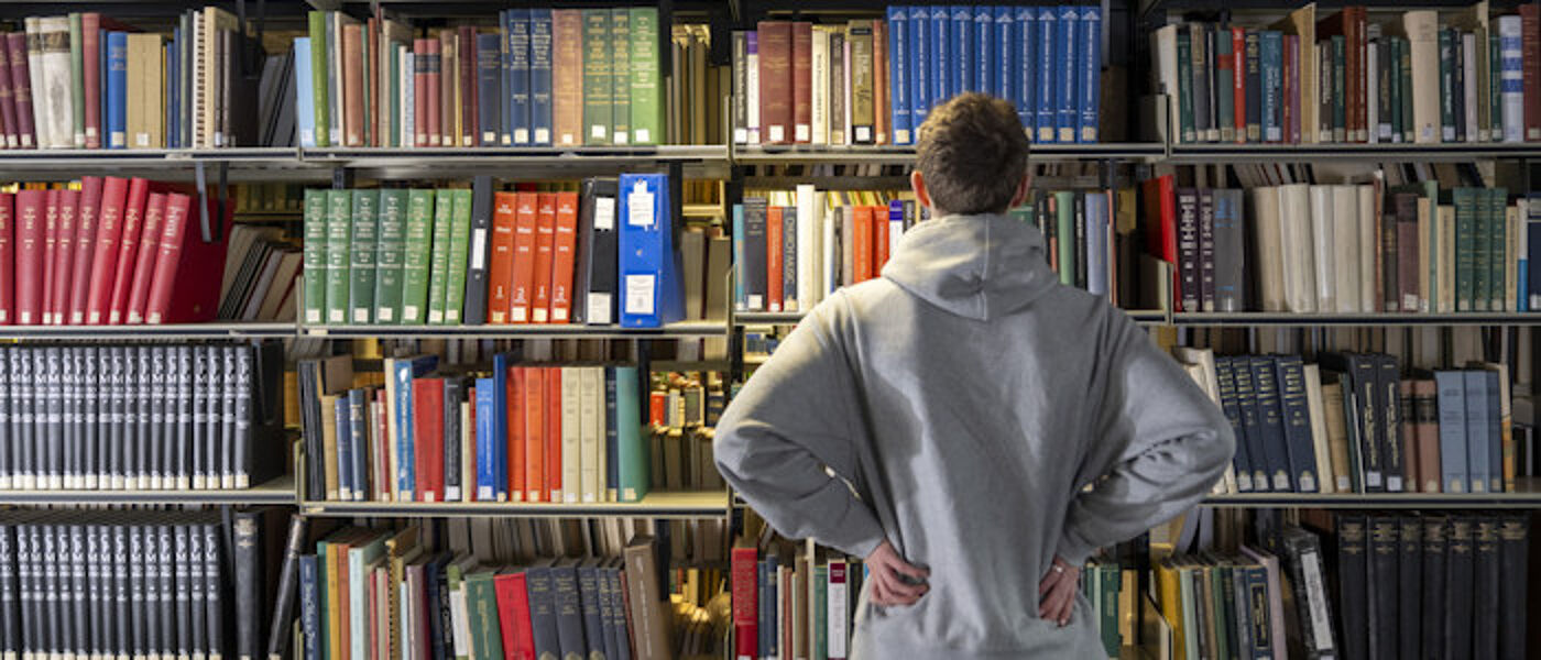 A student faces rows of books on a bookshelf in the library as he contemplates studying