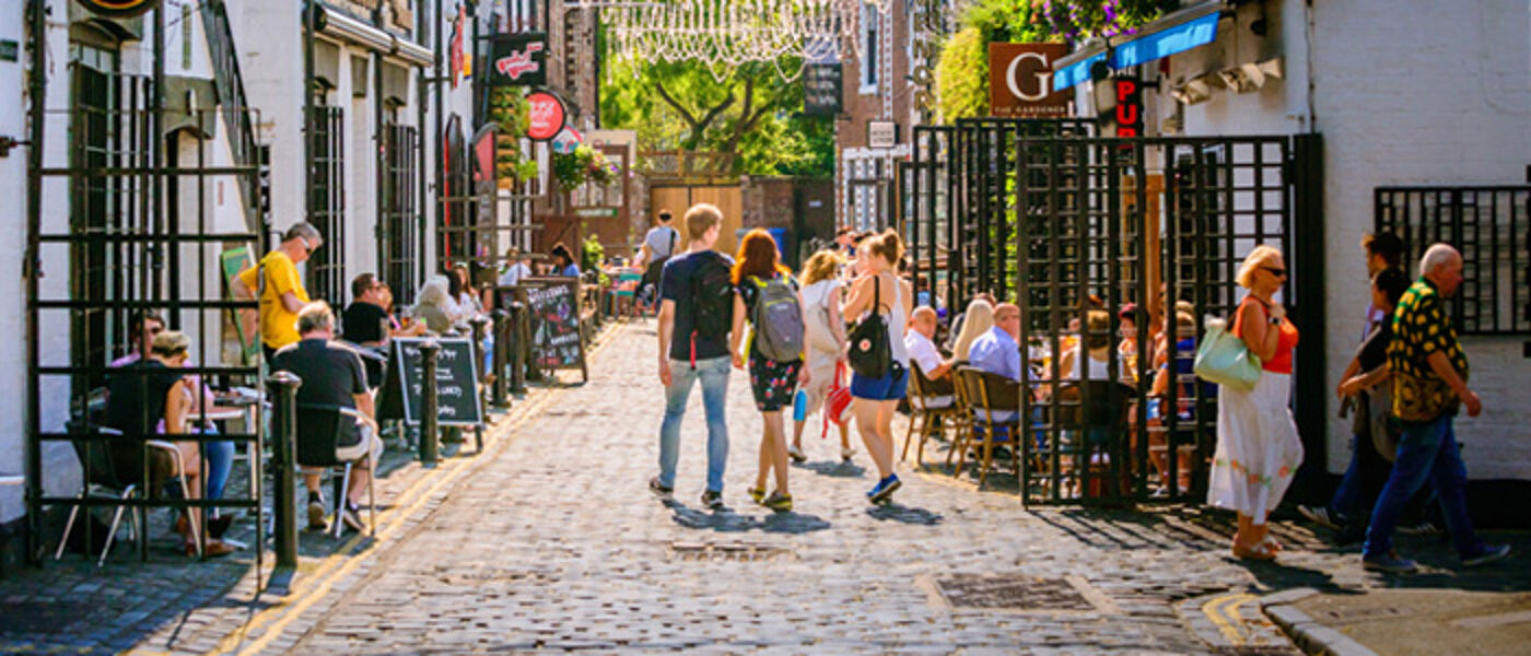 Students walking down Ashton lane, a pretty cobbled street on a sunny day, surrounded by people sitting outside cafes and restaurants eating and drinking, with hanging baskets of flowers and trees in the distance.