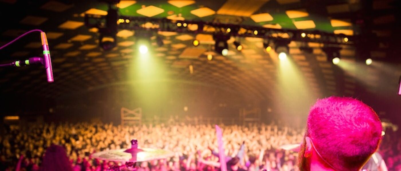 A photograph of a band in front of a large crowd, taken from behind the drummer.