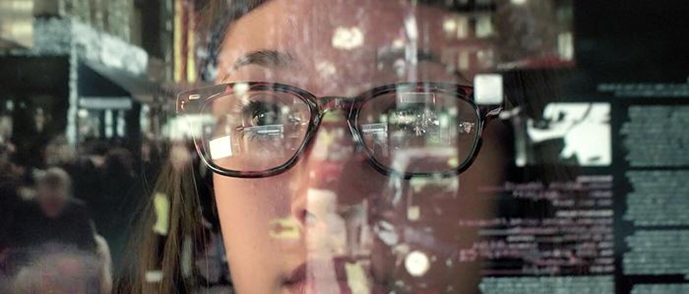 Female looking through glass with reflections of people talkyng and computer screens. 700 pixels