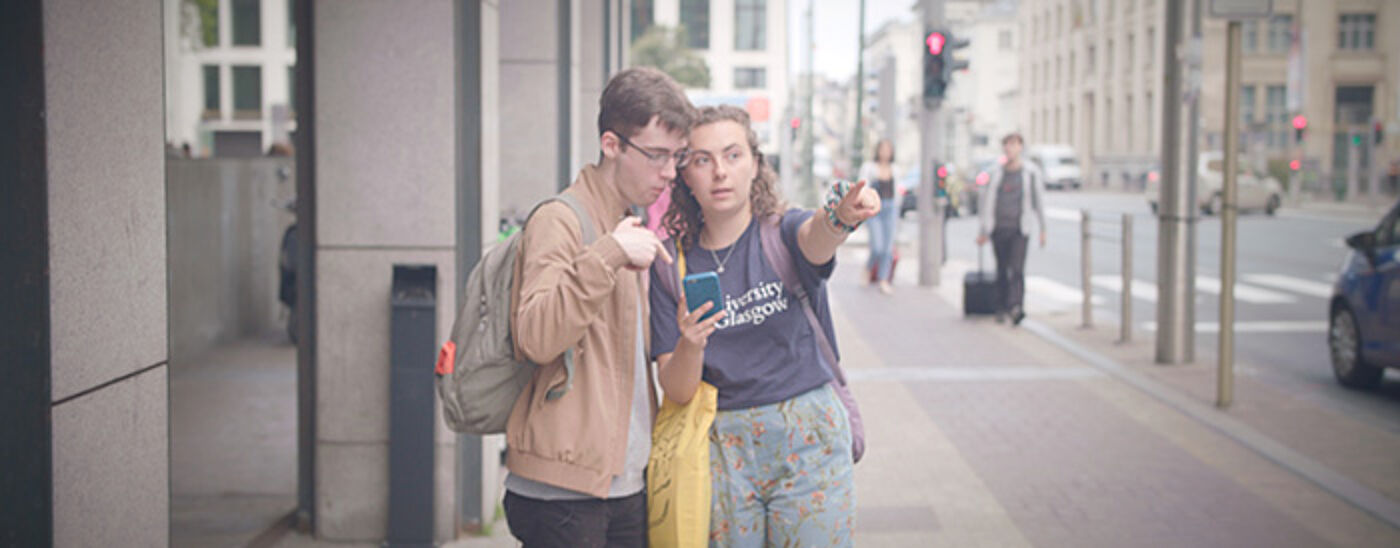 Two students (Ally and Nick) standing in a city street, one pointing ahead, the other looking down at a phone