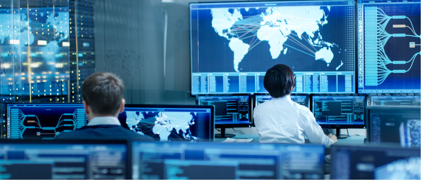 In the System Control Room Operator and Administrator Sitting at Their Workstations with Multiple Displays Showing Graphics and Logistics Information.