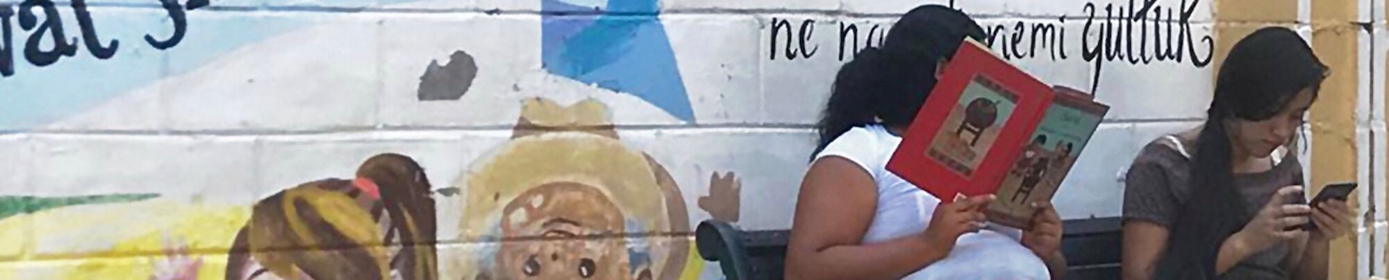 two women reading books on a bench infront of a colourful mural