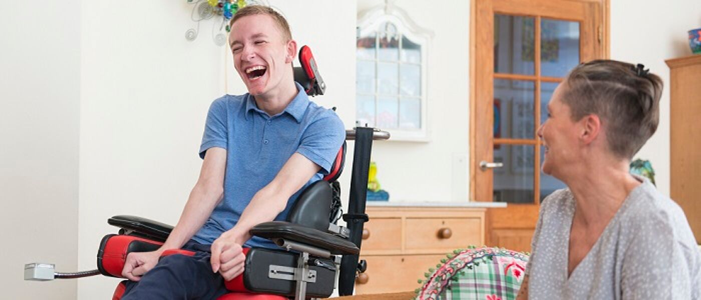 A young man with Amyotrophic lateral sclerosis in a wheelchair in a domestic lounge; he is laughing and sharing a joke with his mpother who is seated on a sofa next to him. Source: iStockPhoto https://www.istockphoto.com/photo/happy-young-als-patient-with-his-mom-gm603871162-103683657