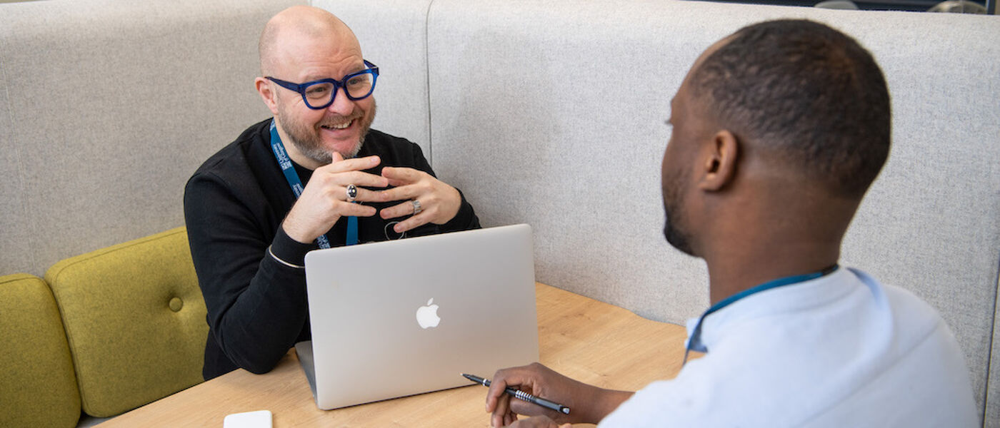 A man with a laptop at at table talking with another man whose face cannot be seen who is making notes with pen on paper