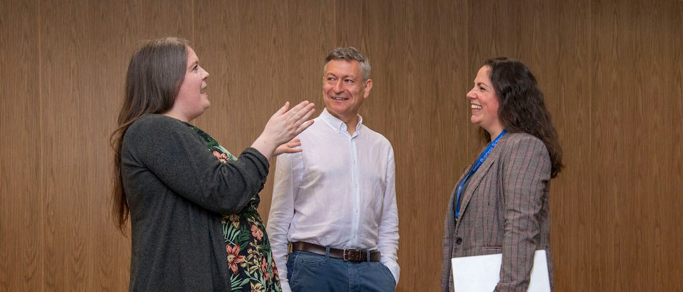 A woman gesturing as she speaks to a man and woman who are smiling as they listen. They are all standing up 