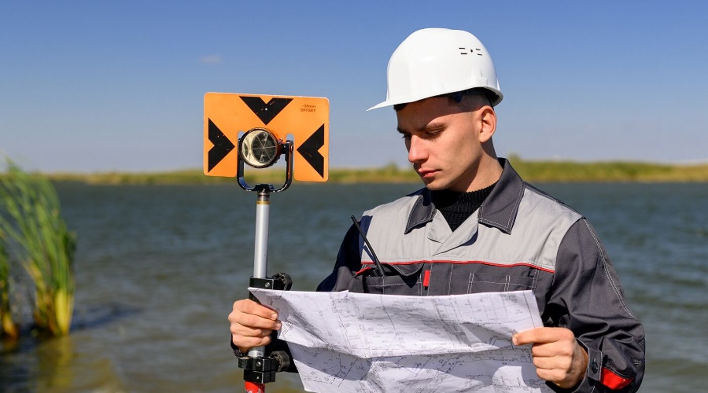 A young man wearing a white hard hat and holding surveying equipment